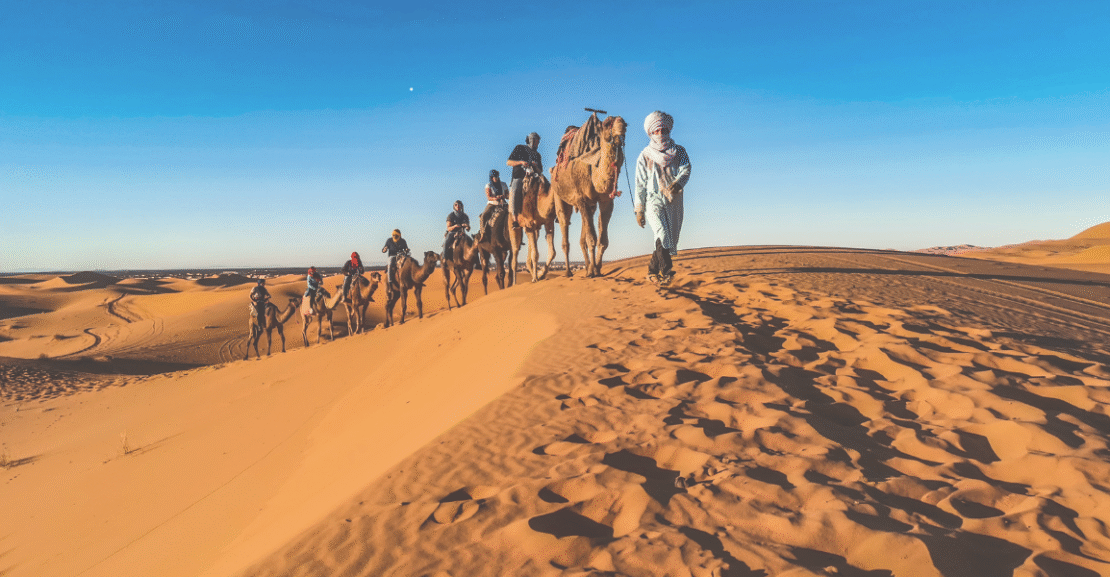 Camel tour in the Merzouga desert, Morocco, with travelers riding through golden sand dunes at sunset.
