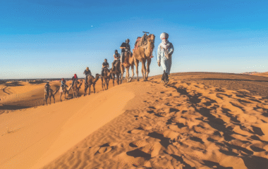 Camel tour in the Merzouga desert, Morocco, with travelers riding through golden sand dunes at sunset.